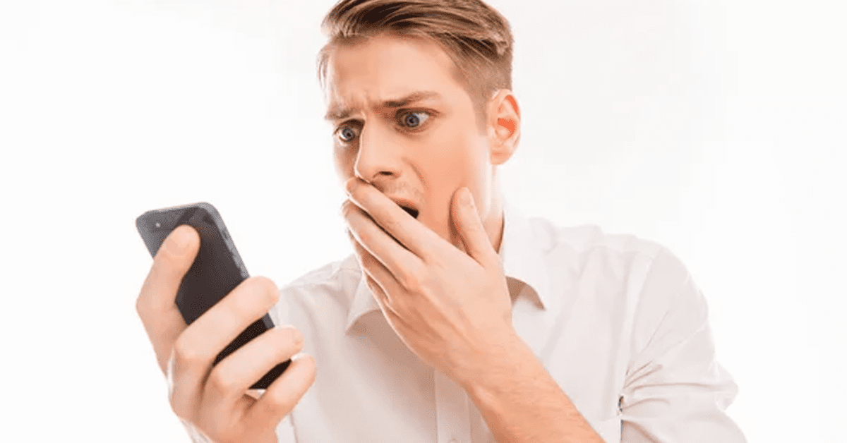 blond guy in a white shirt holding his phone surprised by what he sees