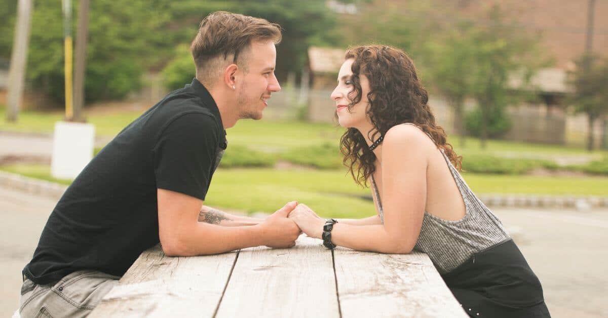 Man and woman sitting facing each other at a picnic table, smiling and holding hands.